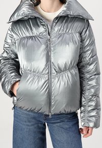 Metallic silver puffer jacket with a high collar, elasticized cuffs, and a cinched waist, featuring a front zipper and side pockets.