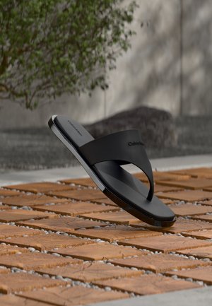 Schwarze Calvin Klein Zehensandale, die über Holzlatten mit kleinen Steinen im Freien schwebt, unscharfer Hintergrund mit Grünpflanzen und Betonwand.