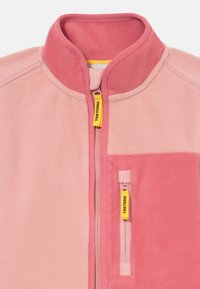 Giacca in pile in diverse tonalità di rosa, con collo alto, frontale con cerniera e tasca sul petto con tirante giallo.