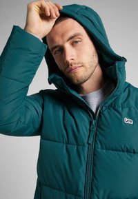 Turkusowa kurtka puchowa z zamkiem błyskawicznym z przodu, wysokim kołnierzem i regulowaną kapturem. Posiada przeszywane panele i małe haftowane logo na klatce piersiowej.