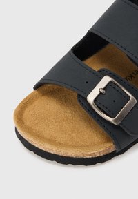 Μαύρο σανδάλι από δερματίνη με φαρδιά λουράκι και ασημένια αγκράφα. Μαλακό καφέ εσωτερικό από σουέντ και εξωτερική σόλα από υφή μαύρου καουτσούκ.