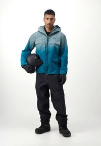 Μπλε-πράσινο και μαύρο μπουφάν για σκι με βαθμιαίο σχέδιο, φερμουάρ μπροστά, δύο τσέπες στο στήθος, σε συνδυασμό με μαύρο παντελόνι για σκι και μπότες. Άτομο κρατάει κράνος.