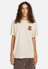 Mladá osoba s dlouhými kudrnatými vlasy v béžovém tričku "Ride the Rails" a tmavě hnědých kalhotách, stojící na jednobarevném pozadí.