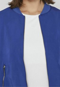Chaqueta bomber azul con una tela suave, cuello acanalado y cremallera dorada. Camisa blanca debajo, mostrando un diseño limpio.