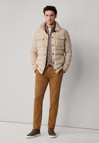 Chaqueta acolchada beige con un alto cuello, bolsillos frontales y botones negros, combinada con pantalones marrones y zapatillas de deporte verde oscuro. Diseño simple.