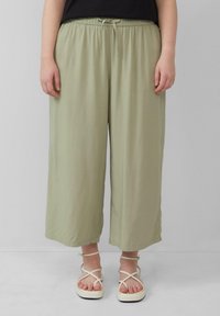 Un pantalon large vert clair présente une taille élastique avec un cordon de serrage. Porté avec des sandales blanches à lanières. Texture du tissu douce et fluide.