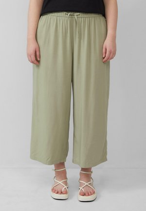 Un pantalon large vert clair présente une taille élastique avec un cordon de serrage. Porté avec des sandales blanches à lanières. Texture du tissu douce et fluide.