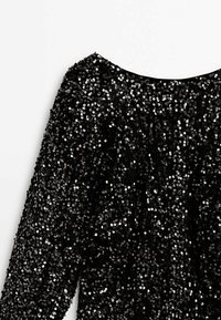 Robe noire à manches longues couverte de sequins noirs brillants, densément serrés, avec un col arrondi sur fond blanc.