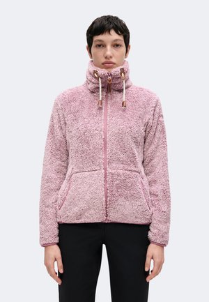 Persona con chaqueta rosa claro de felpa con cremallera, cuello alto y cordones, combinada con pantalones negros, mirando hacia delante sobre un fondo sencillo.