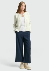 Tânără cu păr lung îmbrăcată într-un cardigan alb cu dungi, top alb fără mâneci, pantaloni largi bleumarin și cizme bej până la glezne, stând cu mâinile în buzunare.