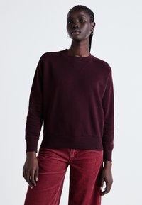 Sweat-shirt bourgogne avec un col rond et des poignets côtelés, présentant un motif texturé. Associé à un pantalon en velours côtelé rouge.