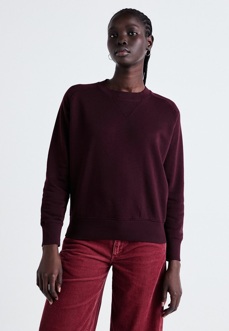 Sweat-shirt bourgogne avec un col rond et des poignets côtelés, présentant un motif texturé. Associé à un pantalon en velours côtelé rouge.