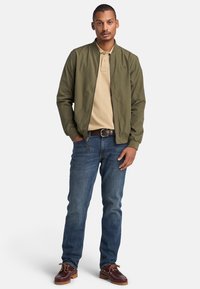 Giubbotto bomber verde oliva con cerniera, polo beige, jeans blu e scarpe di pelle marrone con lacci. Outfit casual classico.