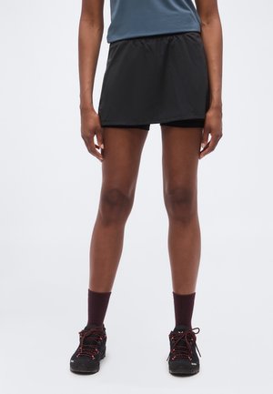 Osoba nosząca czarny sportowy skort, ciemne skarpety i czarne buty do biegania z czerwonymi sznurowadłami, stojąca na jednolitym tle.