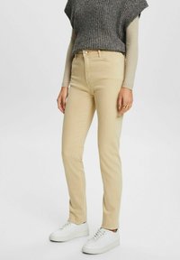 Jeans de un beige claro con un diseño ajustado, que cuentan con cinco bolsillos y una costura sutil, combinados con una blusa de punto gris y zapatillas blancas.