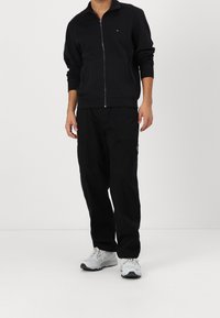Felpa nera con zip, maniche lunghe e tasche frontali, abbinata a pantaloni neri larghi e scarpe da ginnastica bianche.