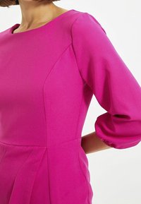 Vestido fucsia con escote redondo, mangas abullonadas hasta el codo, tejido texturizado y detalles plisados en la cintura.
