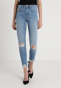 Jean skinny bleu clair avec des déchirures aux genoux, dotés d'une taille haute et d'un léger dégradé. Assortis avec des chaussures à talons hauts noires.