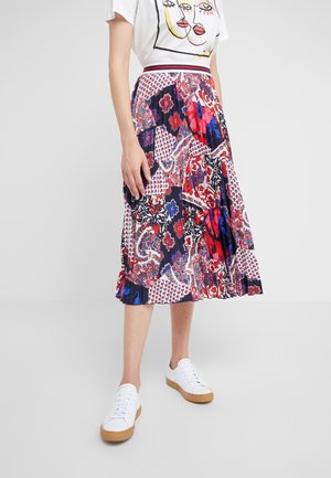 Femme portant une jupe midi plissée à motifs colorés, un t-shirt blanc avec un graphisme et des baskets blanches avec semelles en gomme.