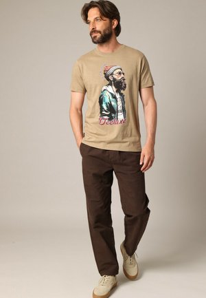 Beige katoenen T-shirt met een illustratie van een man met een baard die een bril draagt, gecombineerd met een broek in bruin en beige sneakers.
