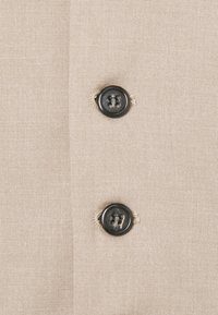 Dva črna gumba s štirimi luknjami, prišita na bež tkanino, postavljena navpično z vidnimi bež šivi okoli luknjic za gumbe.