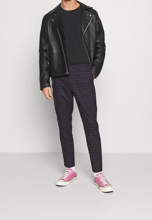Person iført sort læderjakke, sort skjorte, mørke ternede bukser, hvide sokker og pink lærredssneakers, der står mod en ensfarvet baggrund.