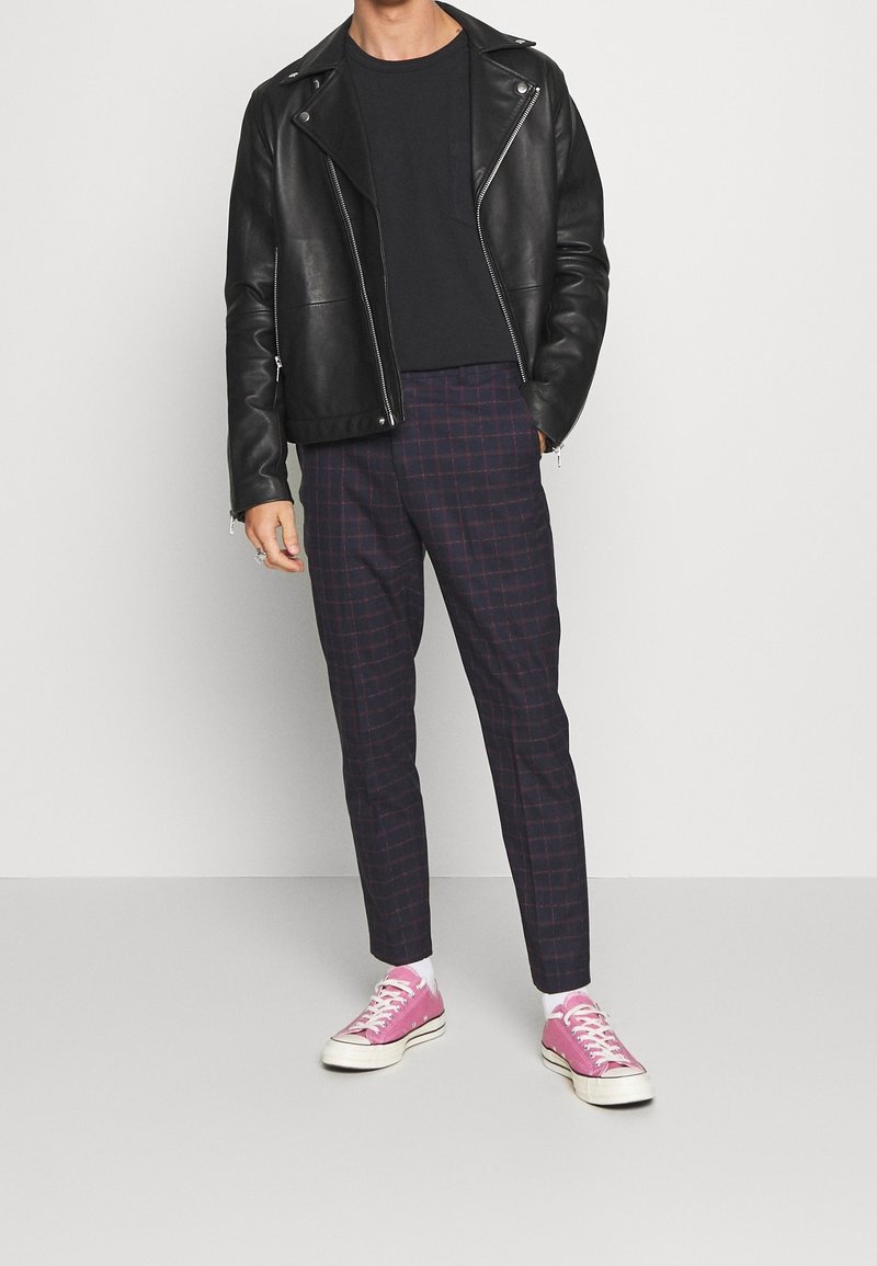 Personne portant une veste en cuir noire, une chemise noire, un pantalon à carreaux foncés, des chaussettes blanches et des baskets en toile roses, debout devant un fond uni.