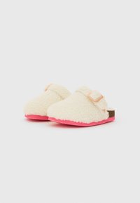 Ett par krämfärgade, pälsiga slip-on-träskor med justerbara remmar och ljusrosa sulor på en vit bakgrund.