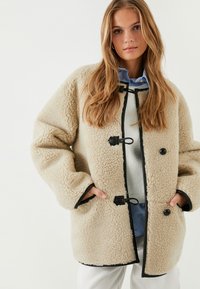 Abrigo de forro polar beige con ribete negro, con un corte relajado, cierres de toggles y bolsillos laterales. Llevado sobre una camisa azul claro.