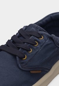 Baskets en toile bleu marine avec des lacets noirs passés à travers des œillets en laiton et une semelle beige, présentées en gros plan.