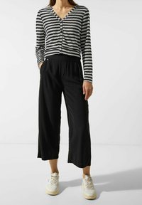 Černé široké kalhoty spárované s dlouhým rukávem topem s černobílým zigzag vzorem, který má výstřih do V a zapínání na knoflíky.