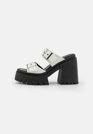 Sandale à talon chunky avec une semelle en caoutchouc noir, une tige en synthétique blanche, deux brides à boucle argentée réglables et une épaisse plateforme.