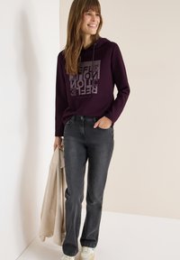 Bordeauxfarbener Kapuzenpullover mit grafischem Druck, schwarze Jeans und heller Sneaker. Der Stoff wirkt weich; die Silhouette ist lässig mit einem entspannten Schnitt.