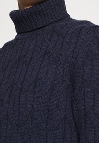 Gros plan d'un pull col roulé en maille côtelée bleu marine porté par une personne, montrant le motif tricoté texturé et le col côtelé.