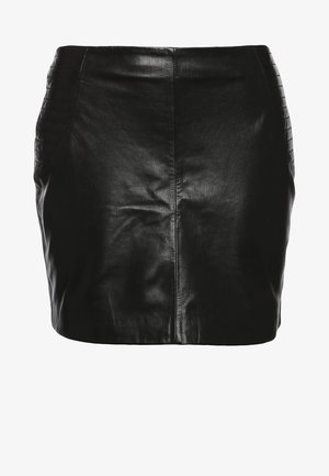 Mujer que lleva una chaqueta y falda de cuero negro, blusa blanca con cuello estampado, medias negras, botines, sosteniendo un bolso de mano en blanco y negro.