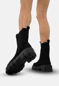 Black ankle boots made of suede, featuring a chunky rubber sole with a tread pattern, a side zipper, and pull tabs for easy wear.