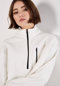 Hvid fleece-pullover med høj krave, der har en sort lynlås foran og en lille lynlåslomme på brystet. Blød tekstur og afslappet pasform.