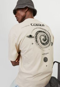 T-shirt couleur crème avec un motif cosmique représentant une galaxie spirale, des planètes et des étoiles en impression noire. Manches courtes, coupe décontractée.