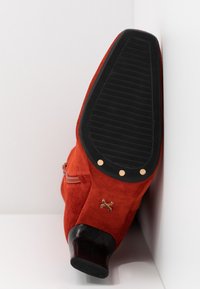 Bottine en daim rouge avec semelle noire, petit talon, clous dorés et fermeture éclair, posée semelle contre une surface blanche d'angle.