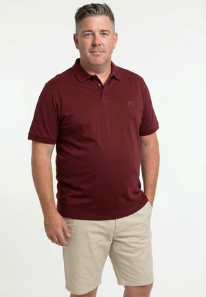 Mand stående med hænderne i lommerne iført en mørkerød polo og beige knælange shorts mod en ensfarvet hvid baggrund.