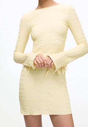 Femme portant une mini-robe à manches longues texturée jaune clair avec un col bateau et un motif de tissu ondulé.