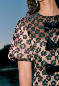 Leopardenmuster-Kleid mit durchsichtigen Puffärmeln, samtartiger Textur und schwarzen Samt-Schleifen als Akzenten auf einem hellbeigen Hintergrund.