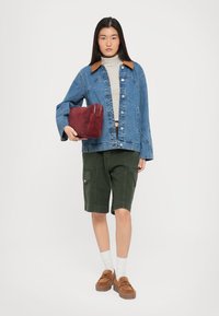 Τζάκετ τζιν με καφέ γιακά, ριγέ σορτς, πράσινα σορτς, καφέ λαστιχένια παπούτσια και κόκκινη θήκη. Το μοντέλο στέκεται μπροστά σε άσπρο φόντο.