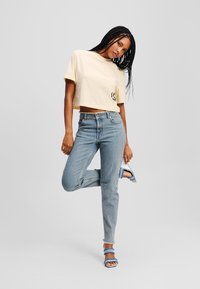 Světle žlutý crop top s černým logem, spárovaný se světle modrými džíny s vysokým pasem a modrými sandály na vysokém podpatku s třásněmi.