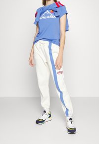 Modré tričko s grafickým motivem hory a textem "RALPH LAUREN", v kombinaci s bílými tepláky se modrým pruhem a červeným logem "POLO". Barevné tenisky.