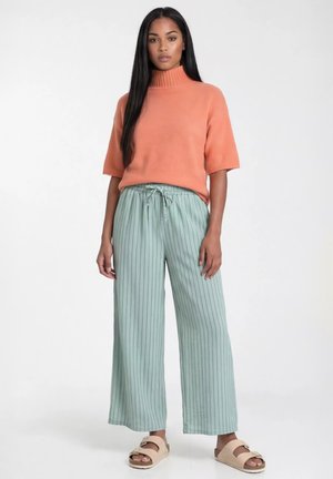 Kobieta stojąca ubrana w koralowy golf z krótkim rękawem, miętowo-zielone szerokie spodnie w prążki oraz beżowe sandały na klamrę na białym tle.