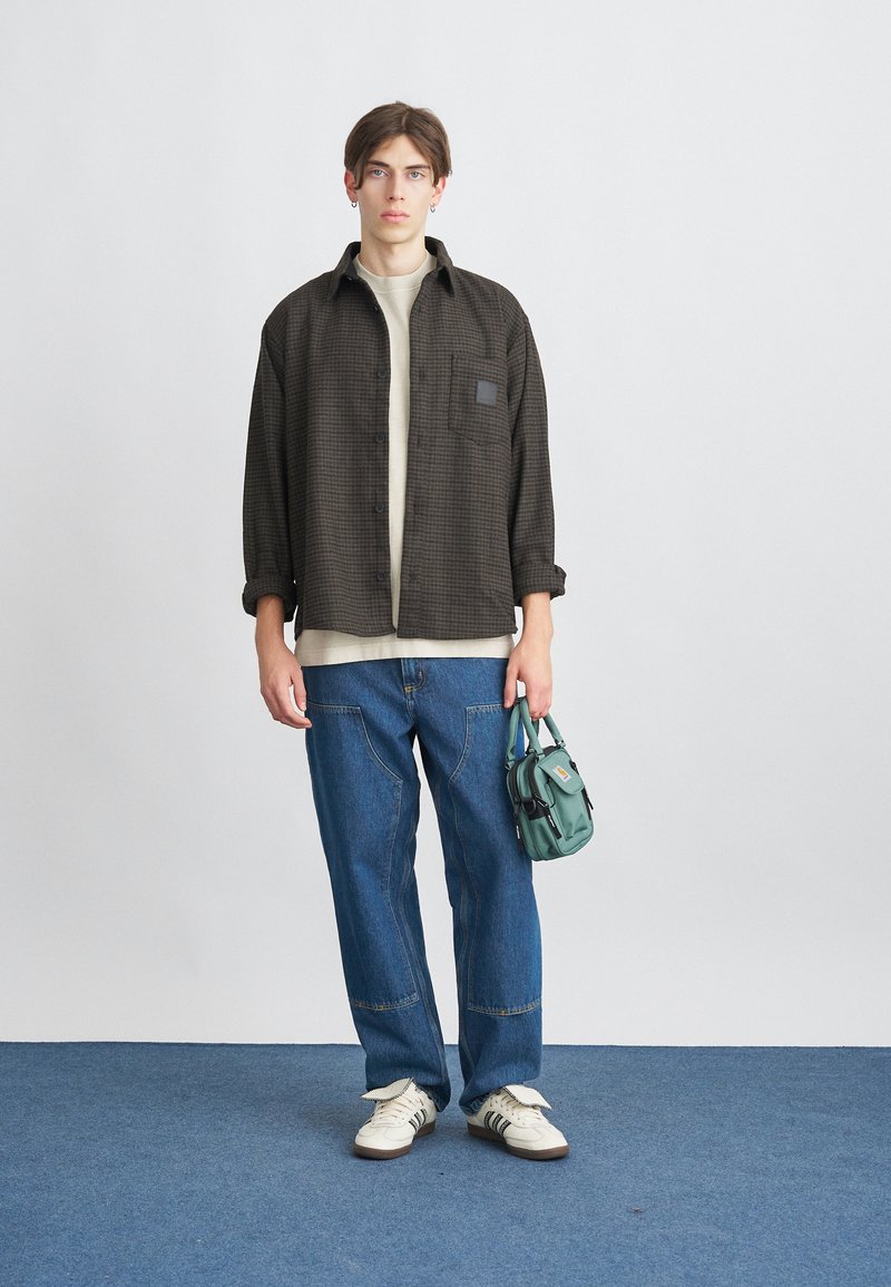 Sort og brun ternet skjorte, beige t-shirt, blå bredbenede jeans og hvide sneakers. Holder en lille teal håndtaske. Neutral baggrund.