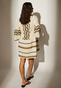 Robe beige avec des motifs en zigzag marron et noir. Manches évasées, ourlet plissé et poches. Fabriquée en tissu texturé.
