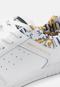 Baskets blanches avec un côté perforé, "Versace Jeans Couture" en texte doré, lacets blancs et doublure intérieure à motifs colorés.