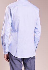 Světle modrá košile s dlouhými rukávy a hladkou texturou, s manžetami na knoflíky a zakřiveným lemem. Obsahuje klasický límec a jemné krejčovské zpracování.